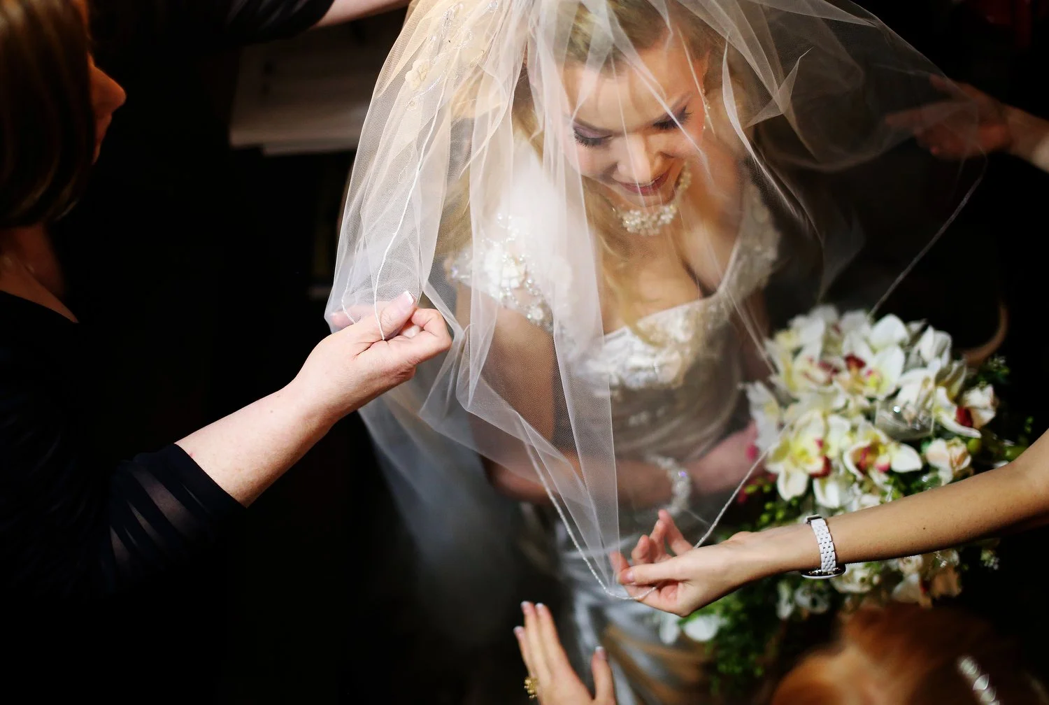 A bride with a veil over her face, smiling, holding a bouquet of flowers, as hands help adjust her veil.