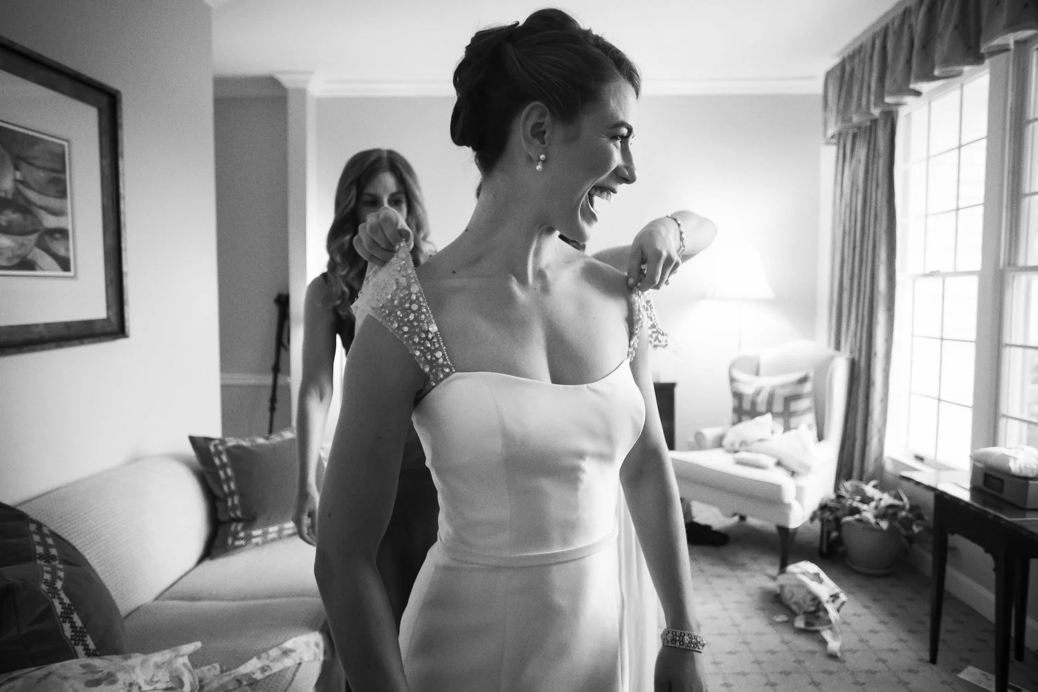 Black and white photo of a woman in a wedding dress laughing as someone helps her adjust the sheer sequined straps of her dress in a well-lit room with huge windows. Another woman is in the background.