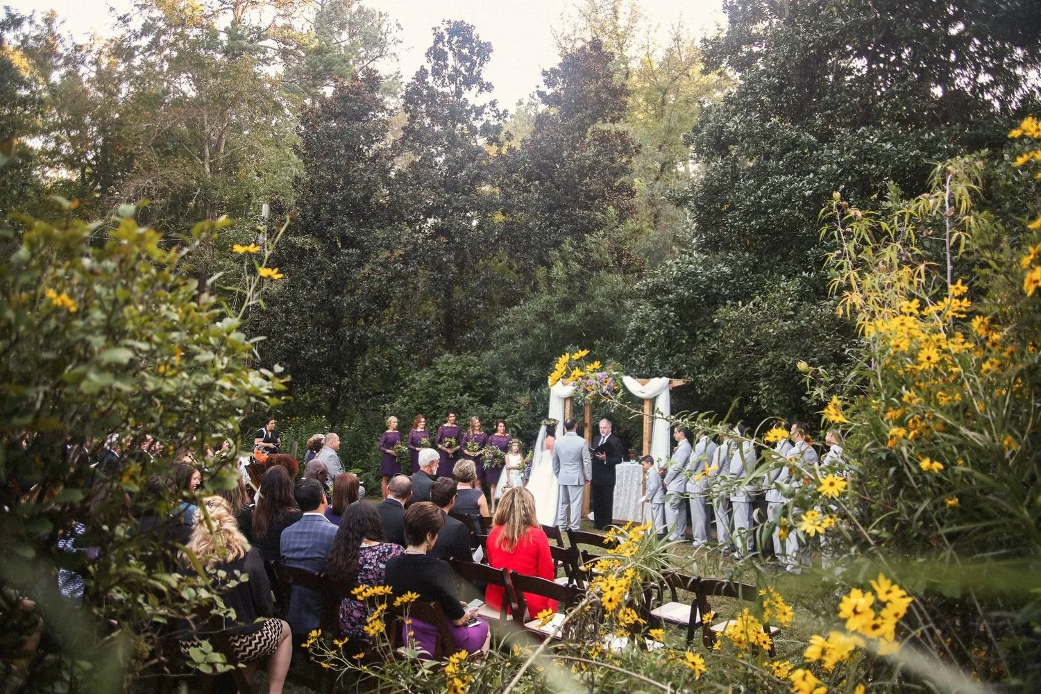 Outdoor wedding ceremony with a couple at the altar, surrounded by guests and wedding party, in a lush garden setting with trees and yellow flowers. The people are small in the distance of this scene setter image.