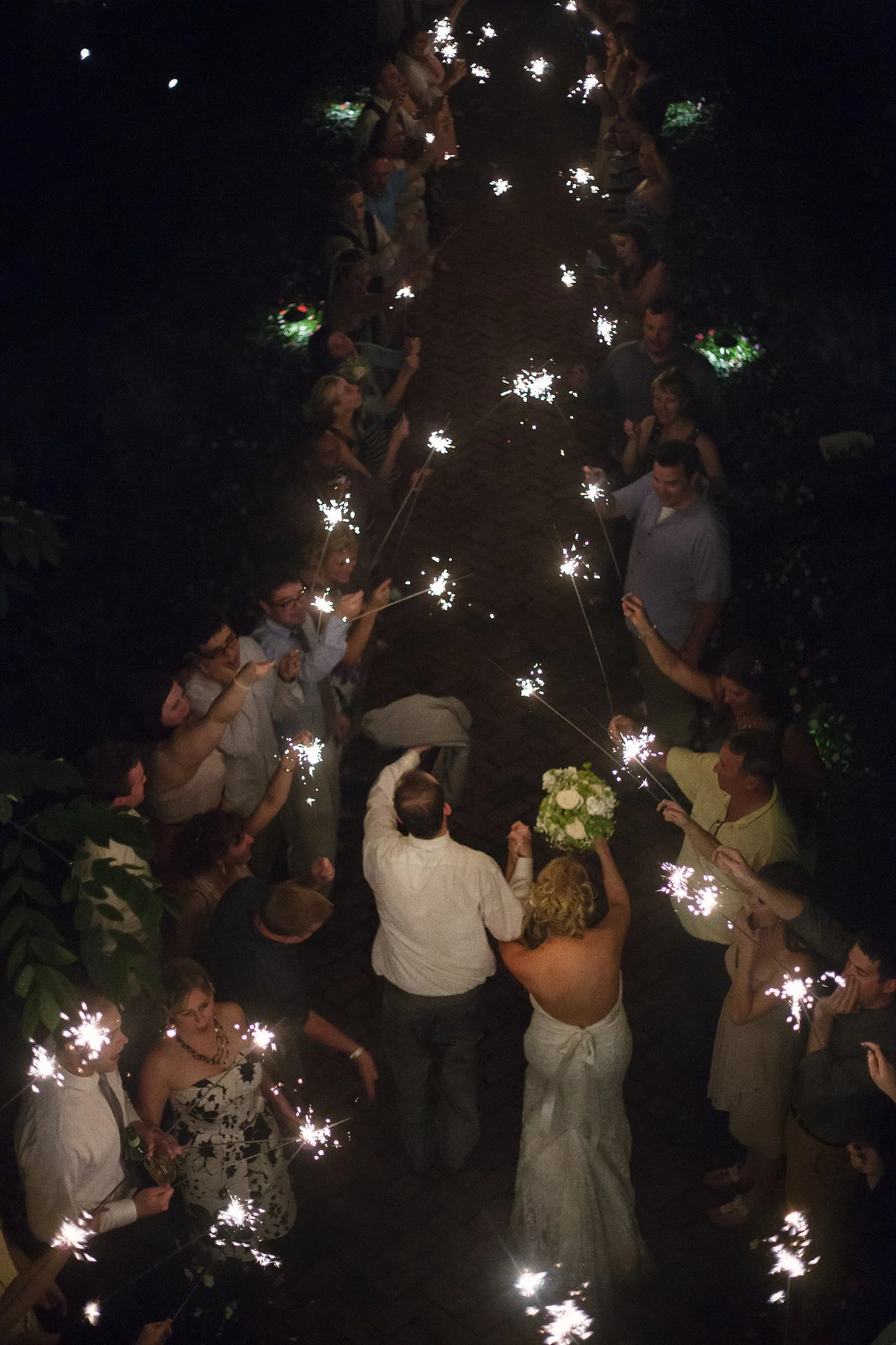 Aerial shot of the back of a bride and groom as they walk through two rows of wedding guests holding sparklers above them in the dark of night. The bride is on the right in a white strapless dress holding her bouquet up above her head. The groom is o