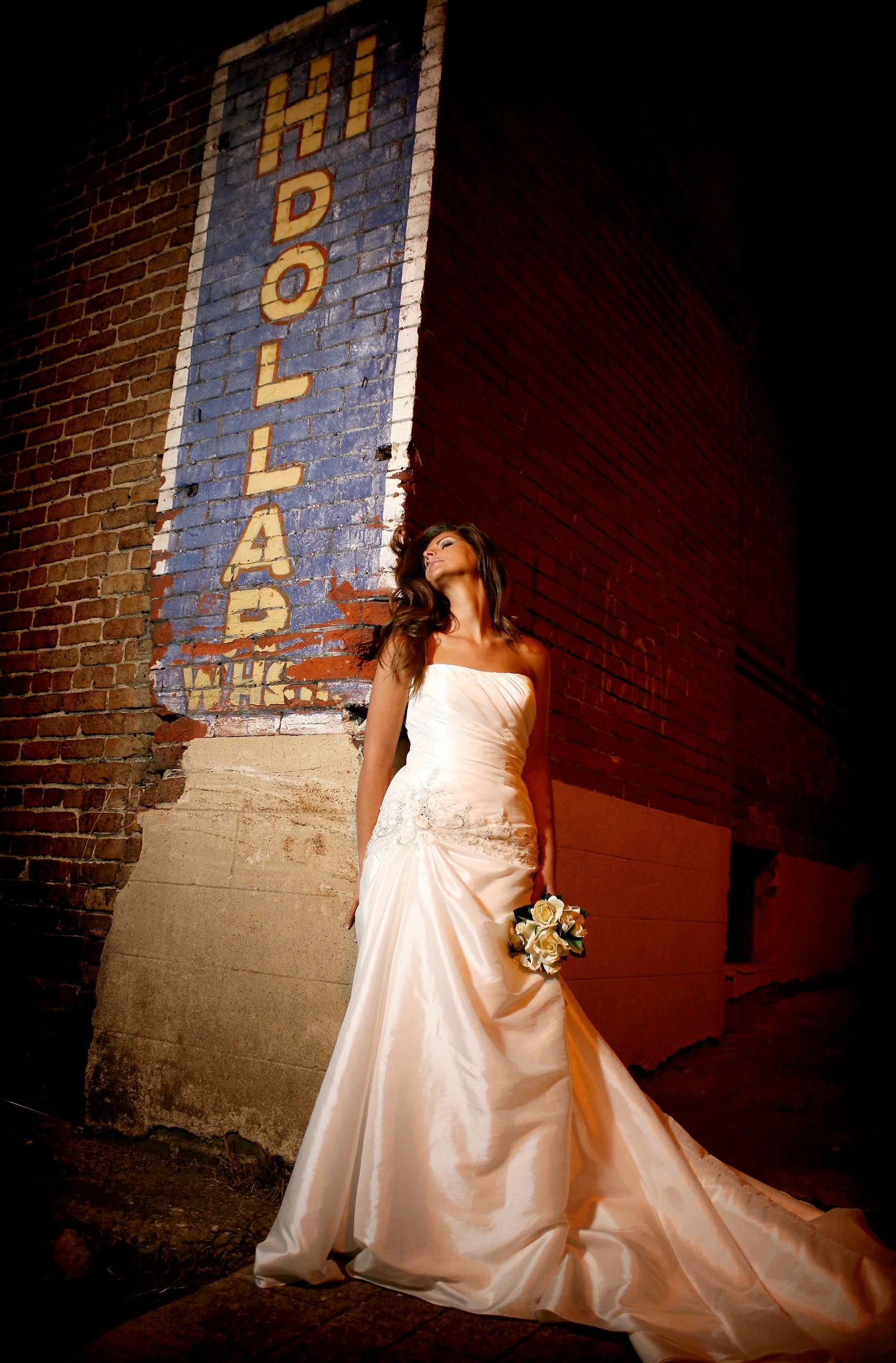 Bridal portrait taken at night. The bride has a long flowy white gown and holds a bouquet of flowers. She arches her head backward as she leans against a brick wall. The words HI DOLLAR are in yellow letters on a vertical blue rectangle of brick with