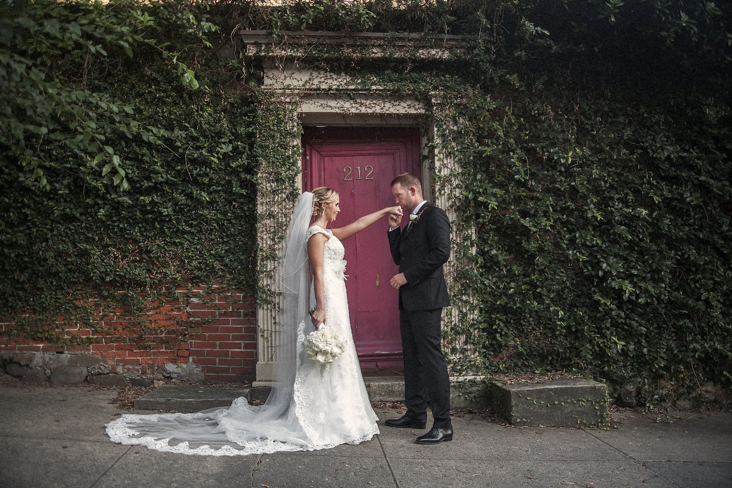 Bride and groom stand in front of a weathered pink door with the number 212, surrounded by ivy-covered walls. The bride, wearing a white lace gown and veil, holds a bouquet of white roses. The groom, in a black suit, kisses the bride's hand.