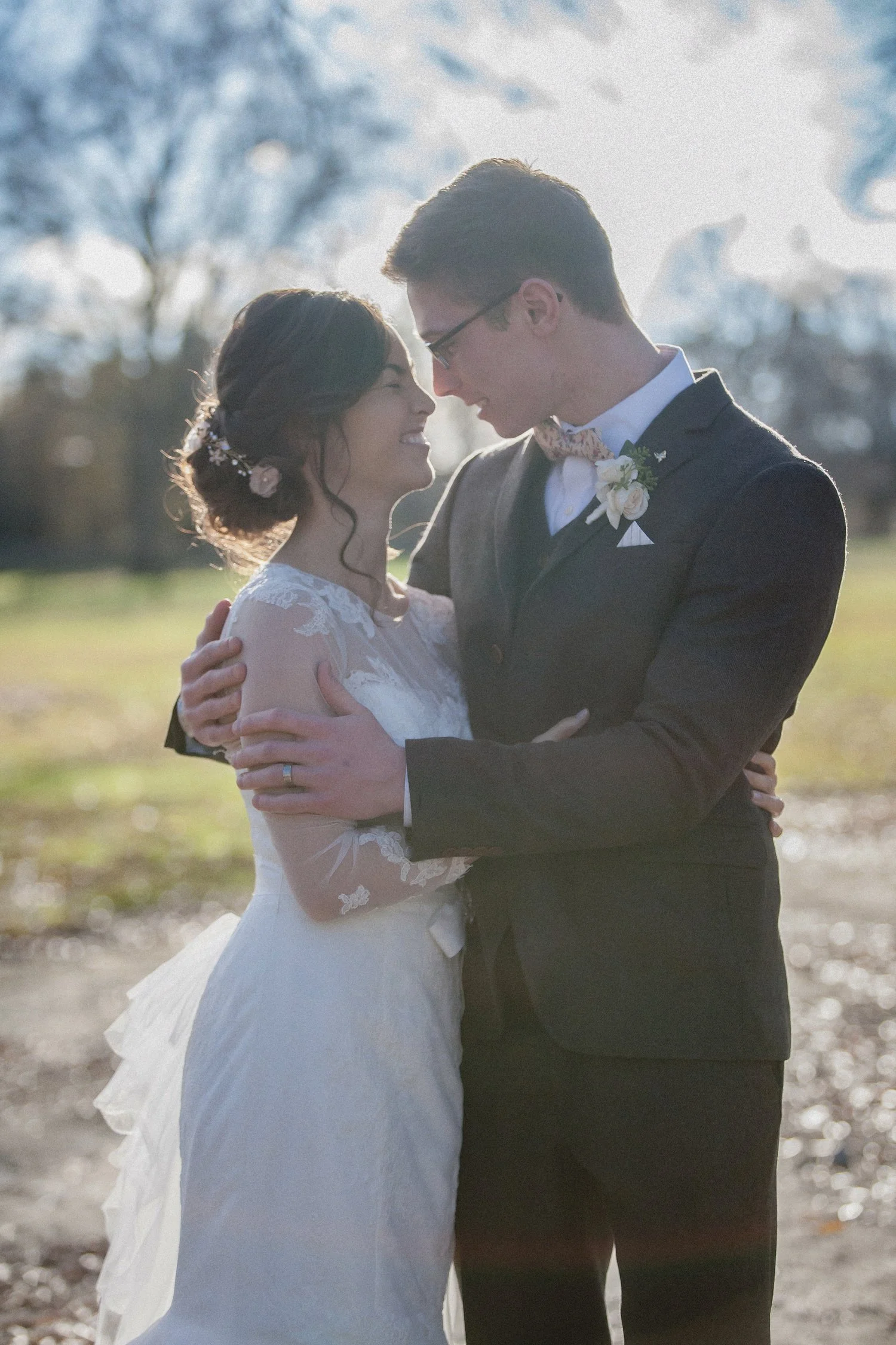 A bride and groom embrace outdoors, smiling at each other, with trees and sunlight in the background.