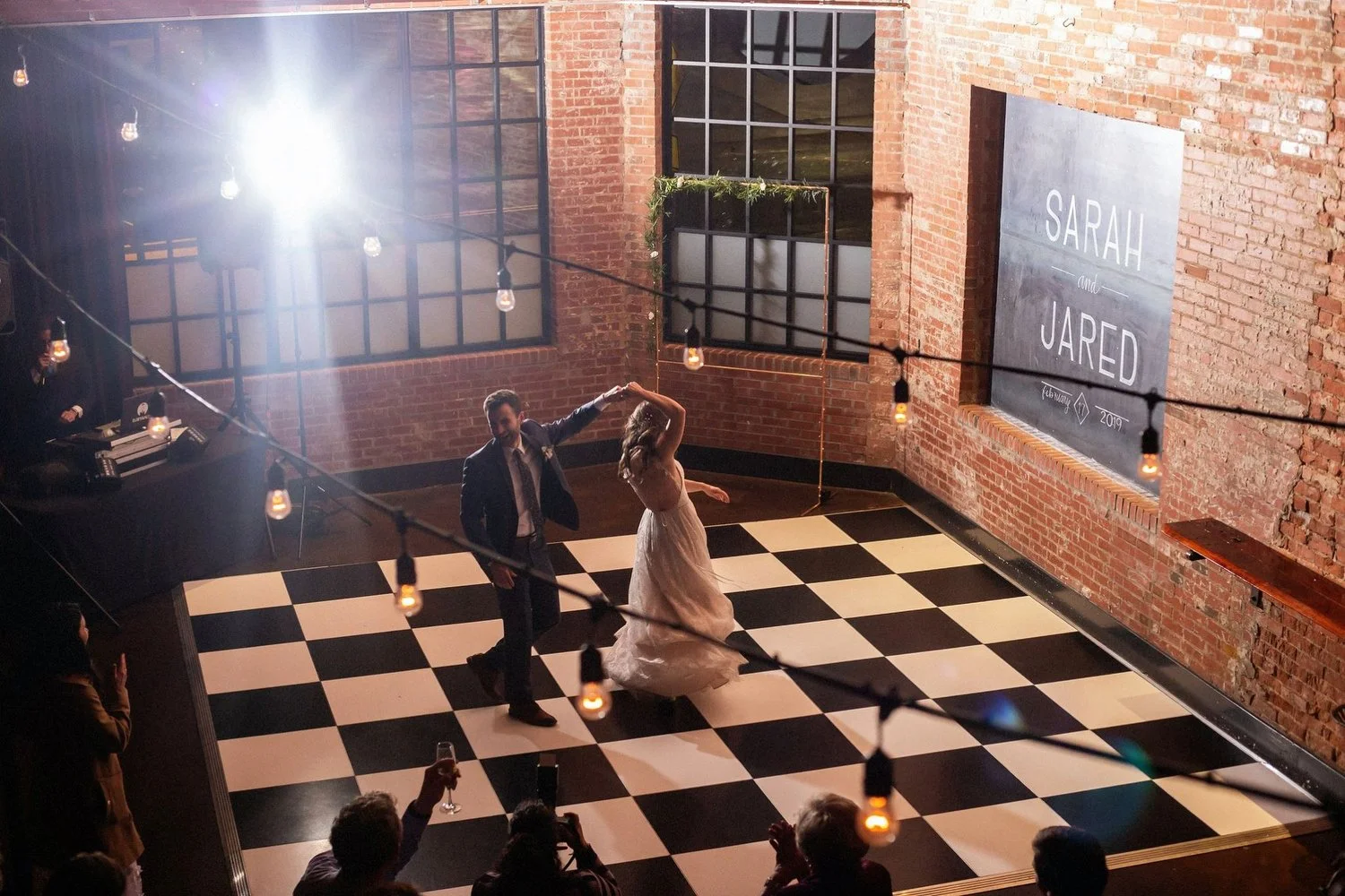 A groom in a black suit with a black tie twirls his bride who is wearing a sleeveless white wedding dress. They are dancing alone on a black-and-white checkered dance floor. There are old industrial windows behind them, twinkle lights above and an ol