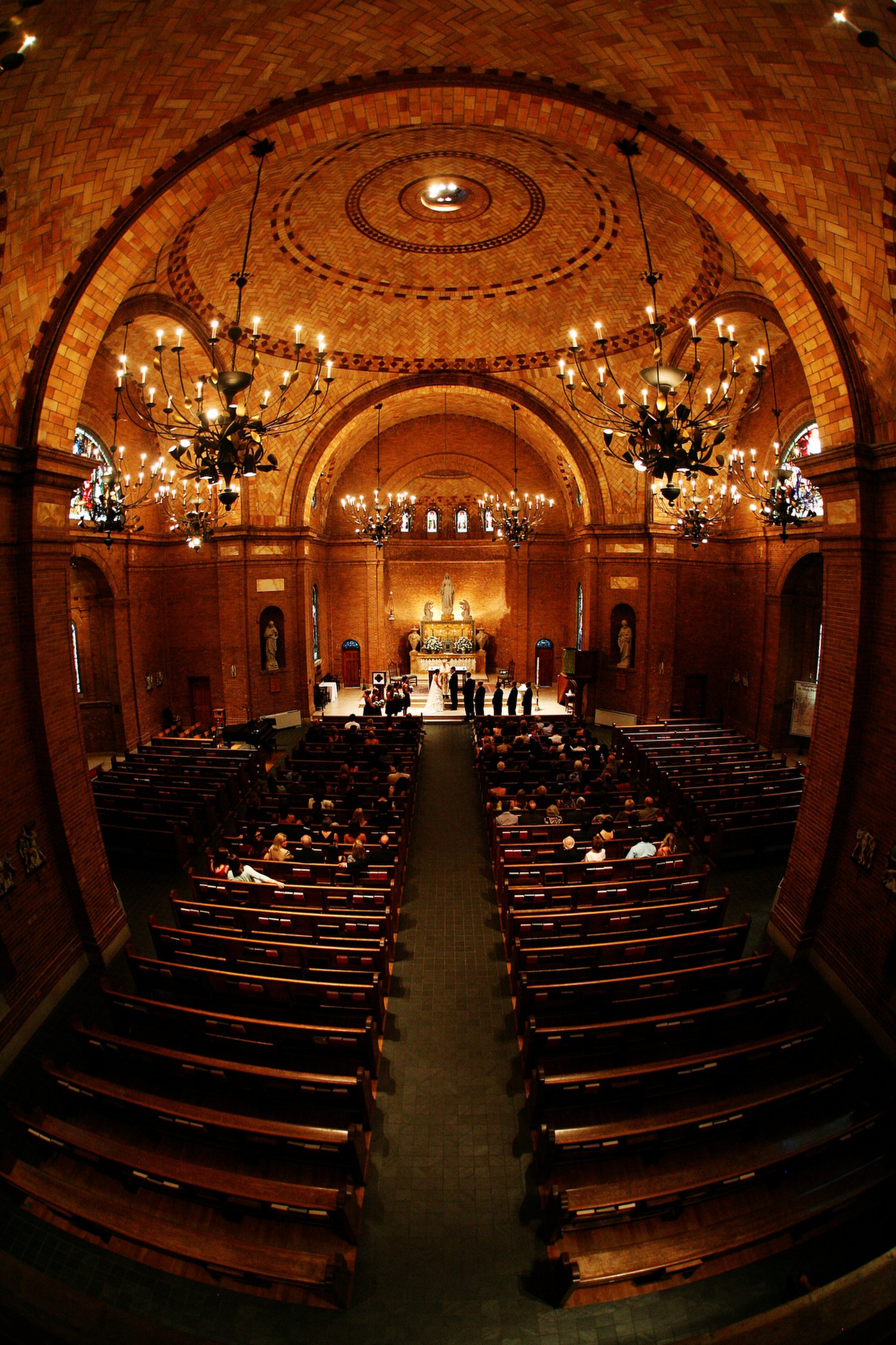 Interior vertical image of a church or cathedral with wooden pews, chandeliers, stained glass windows, and an altar with religious statues. The image has an orange glow as the lights illuminate the curved brick ceiling.