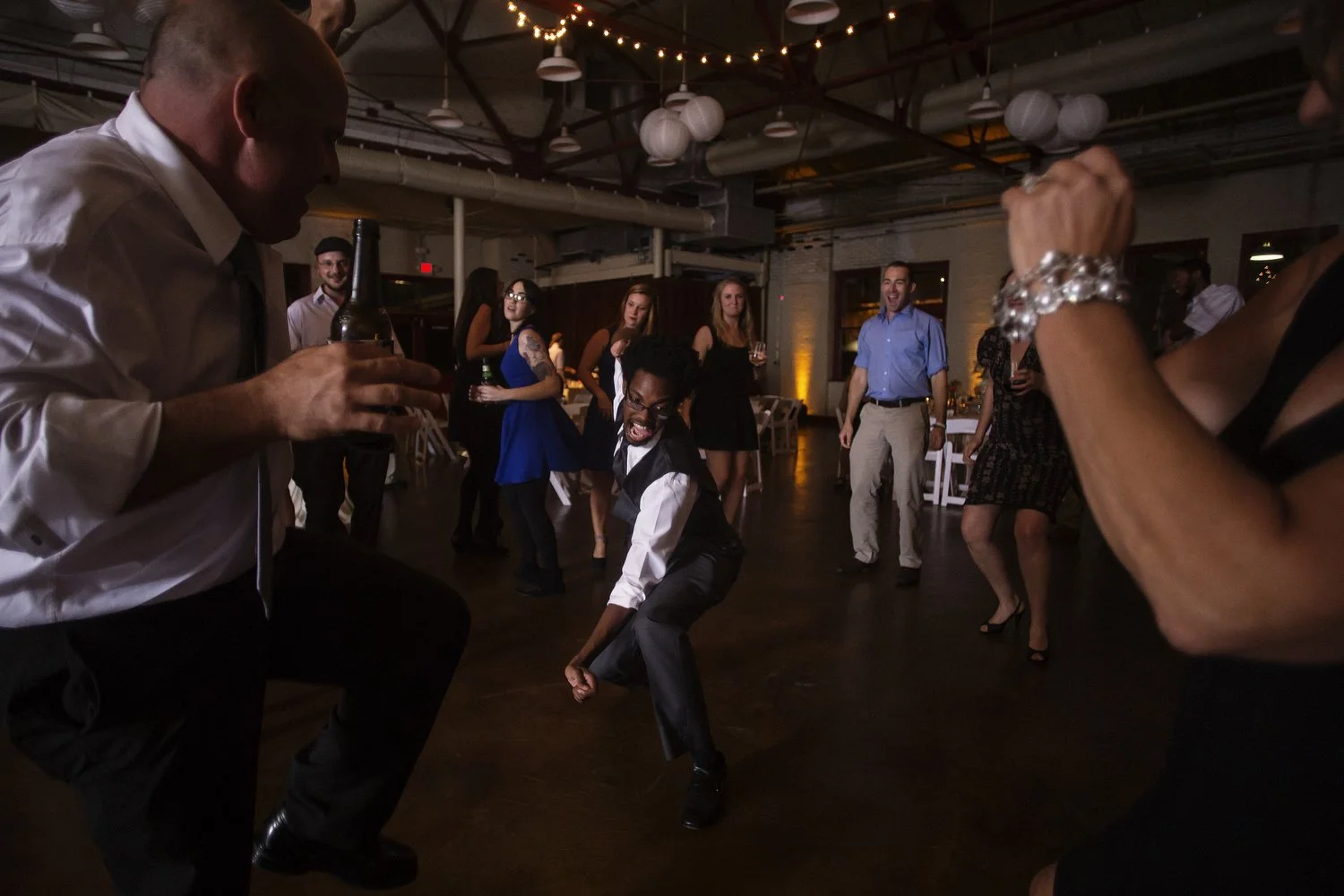 Photo of a dark reception dance floor. One young man wearing a black vest and white button-down shirt kneels in the middle with his arm outstretched doing some sort of dance move. People are all around. There are a few twinkle lights up above.