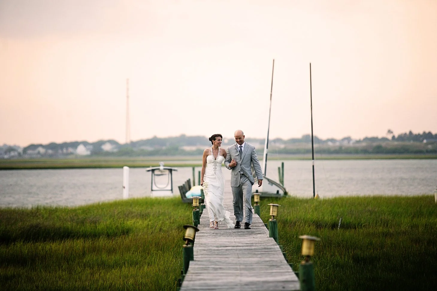 Bride and groom walking and holding hands on a weathered dock near the coast. There is water and a few boats in the distance behind them, and green marshy grass on both sides. They are small in the frame as the image is taken from a distance. He has 