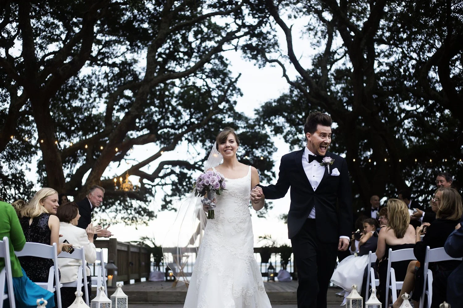 Bride and groom walking hand-in-hand down the aisle at their outdoor wedding ceremony, surrounded by seated guests and large trees in the background. Their wedding ceremony was facing the water. She is wearing a white sleeveless dress and veil and cl