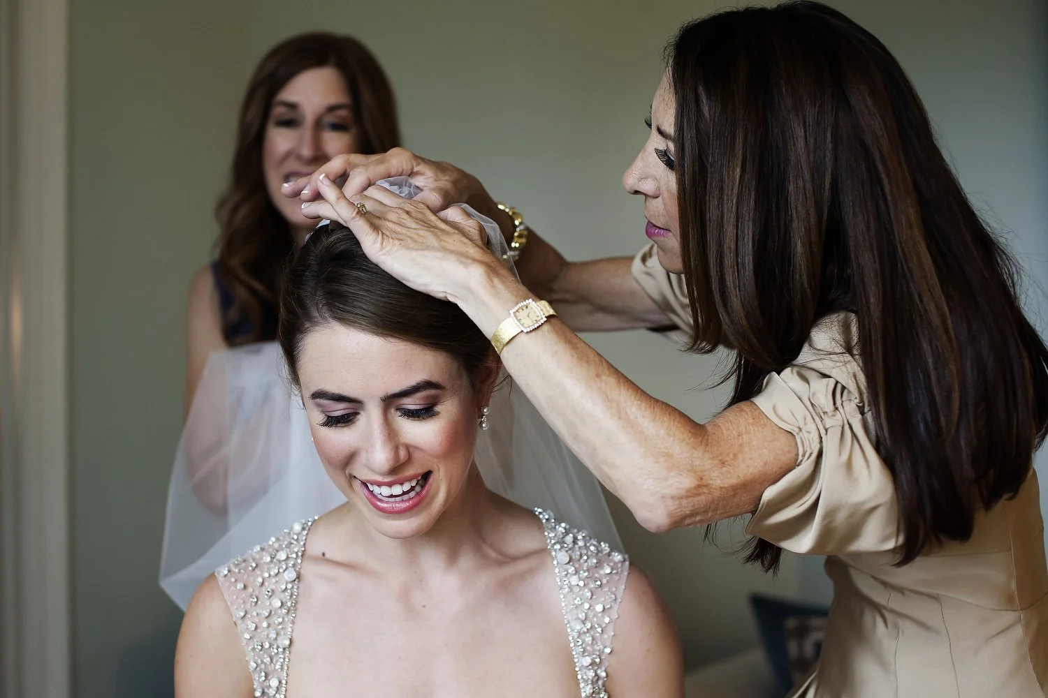 A bride sits and smiles while looking down as her mother adjusts her veil. The straps of her wedding gown are all that's visible and they are sheer and covered with jeweled beads. Her mom's long brown hair is down and she's wearing a tannish-gold col