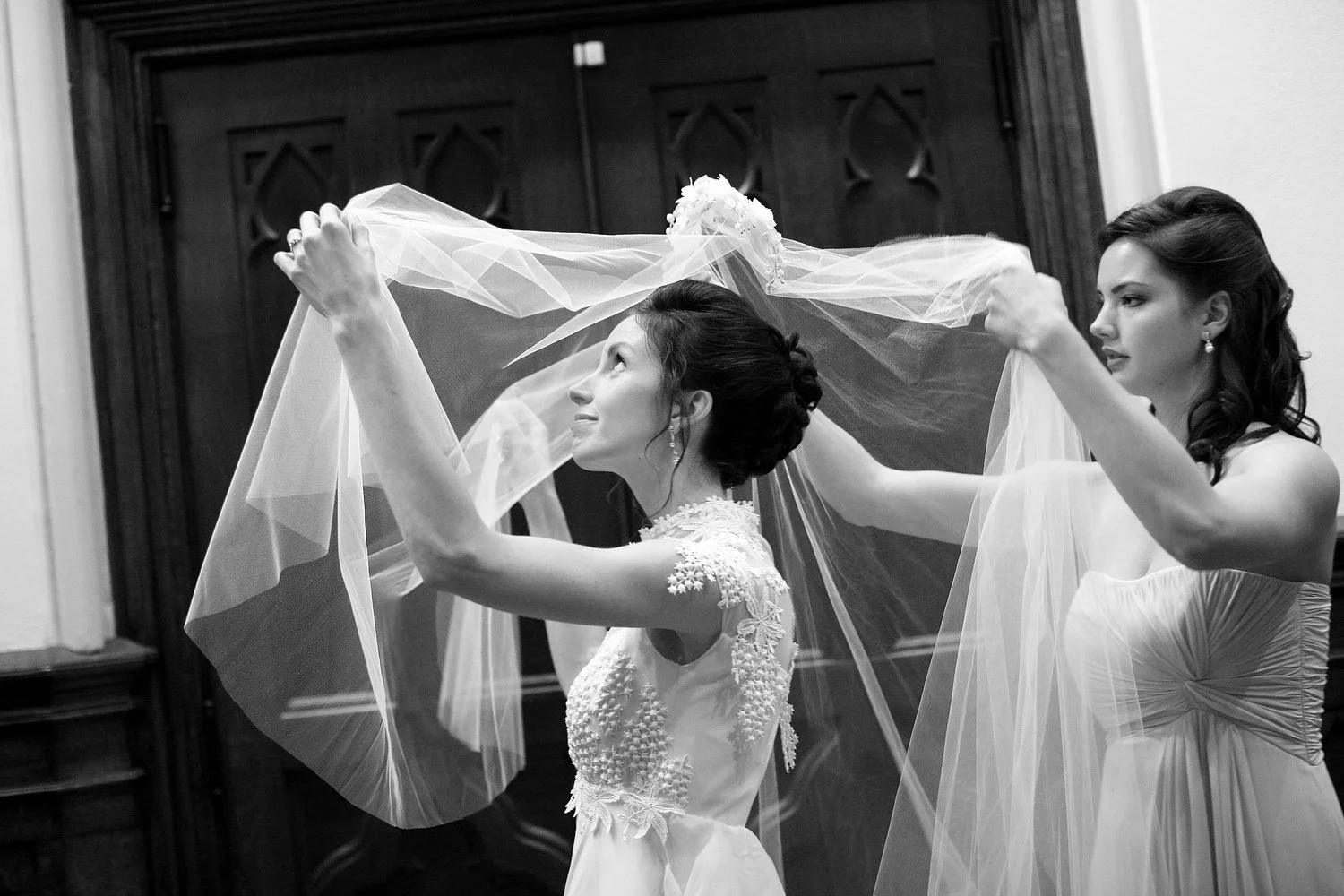 Black and white image of a bride with styled hair and a vintage lace wedding dress standing as she puts her veil over her face, with her bridesmaid assisting her, in front of a dark wooden background.