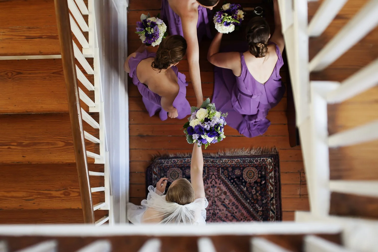 Overhead view of a bride handing a bouquet to a bridesmaid on a wooden staircase during a wedding ceremony, with other bridesmaids in purple dresses holding bouquets.