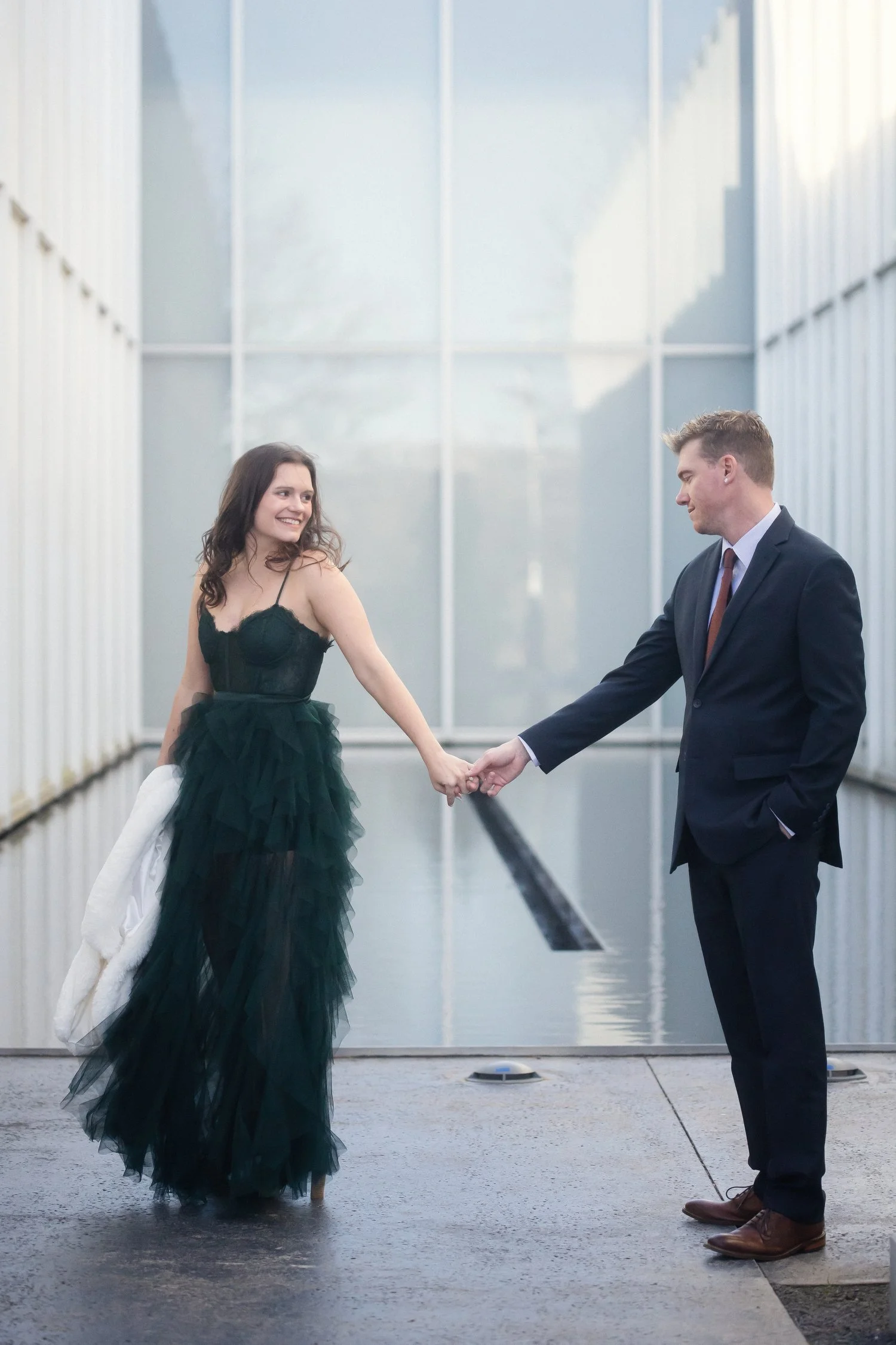 A couple in formal wear stands apart and reaches toward each other to hold hands. There are huge glass windows behind them and on both sides of them as if they are in an outdoor courtyard in between building walls. Sh has on a dark green dress with s