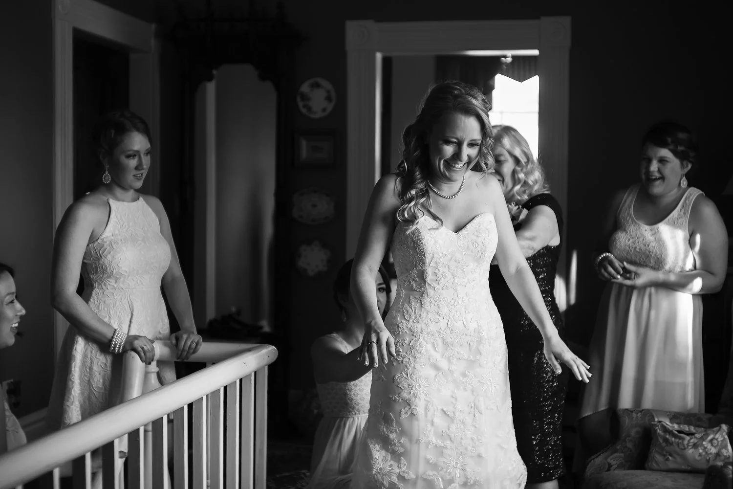 A bride in a white wedding dress smiling, surrounded by women at the landing at the top of a wooden staircase. There's a room with an open door and a window behind her.