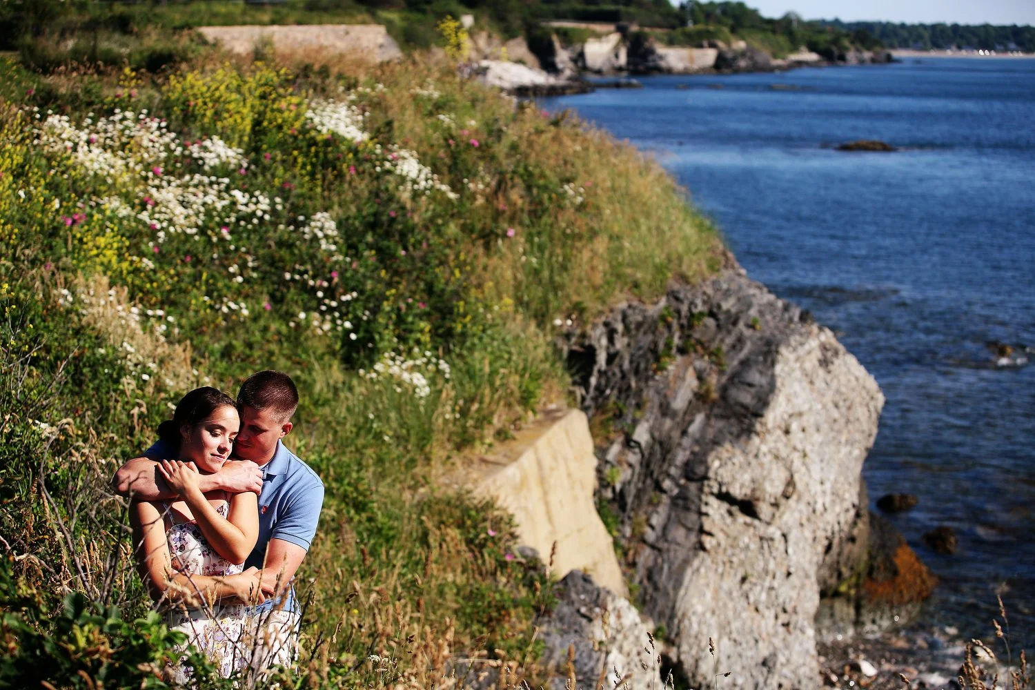 A couple stands in an overgrown field with wildflowers on the side of a rocky cliff next to the water. He has on a Carolina blue short-sleeved shirt with khaki pants and has one arm around her waist and the other around her chest and shoulders. She i