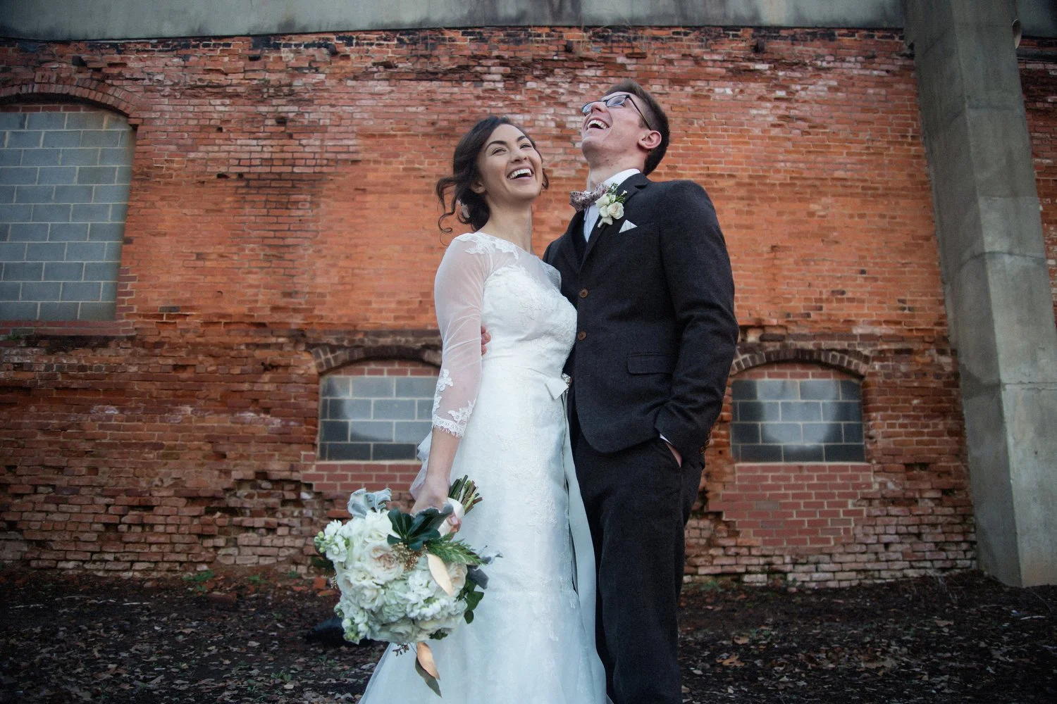 A newlywed couple, the bride in a white wedding dress with sheer sleeves holding a bouquet, and the groom in a dark suit with glasses, happily laughing together outdoors in front of a weathered brick wall.