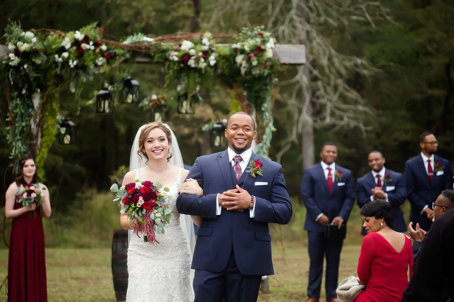 A newlywed couple walking down the aisle at an outdoor wedding, with guests and a decorated arch in the background. The bride is holding a bouquet and is in a white wedding dress, while the groom is wearing a navy suit with a red boutonniere.