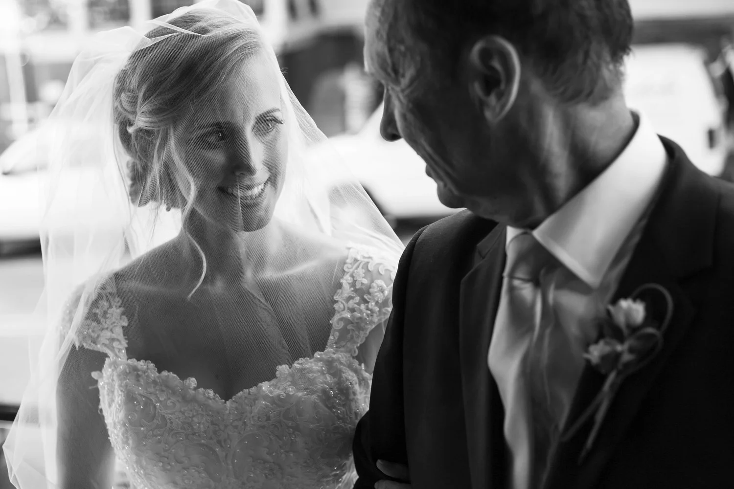 A black and white photo of a bride in a wedding dress and sheer veil over her face smiling at an older man in a suit, possibly her father.