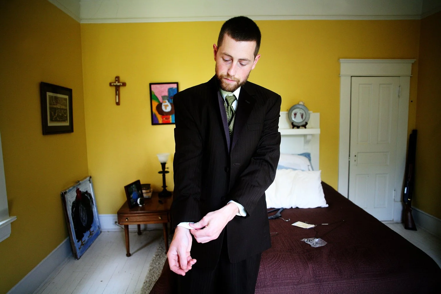 A man in a black suit adjusting his shirt cuff in a bedroom with bright yellow walls, decorated with a crucifix and colorful art, a wooden nightstand, and a bed with white pillows. This is at Elodie Farms.