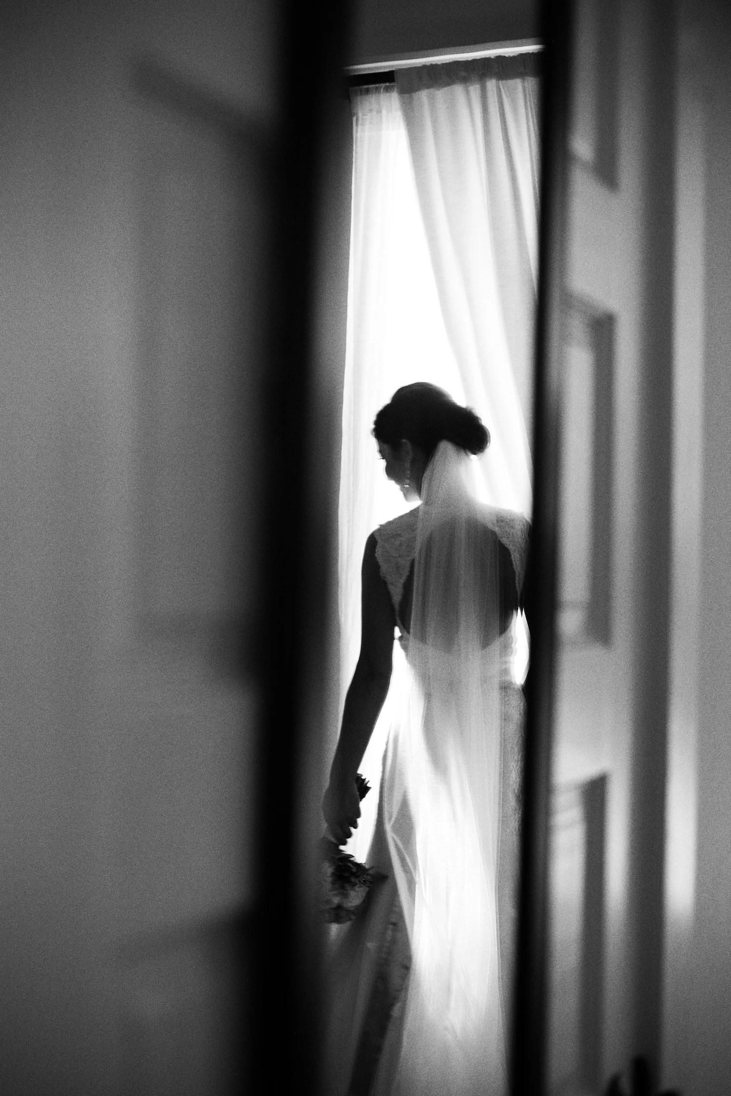 Black and white photo of a reflection of a bride in a wedding dress and veil, silhouetted near a window with sheer lace curtains.