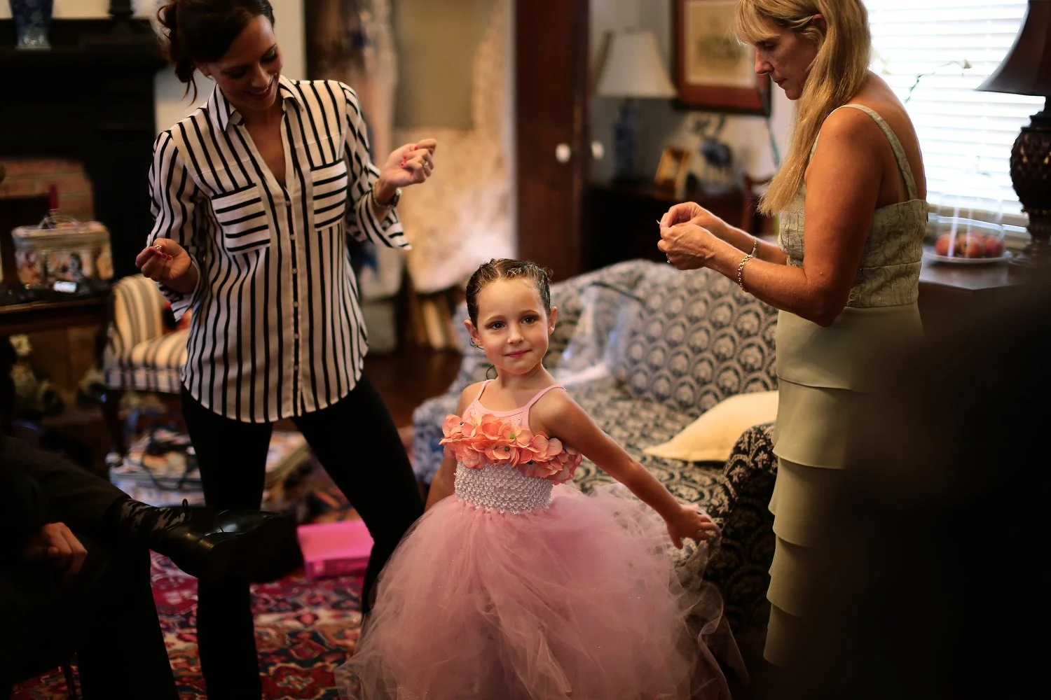 A young girl in a pink tutu dress with flower embellishments is dancing or posing while two women watch and smile in a cozy, warmly lit room. One woman has a black and white striped silk shirt. The other has a mint green dress with spaghetti straps.