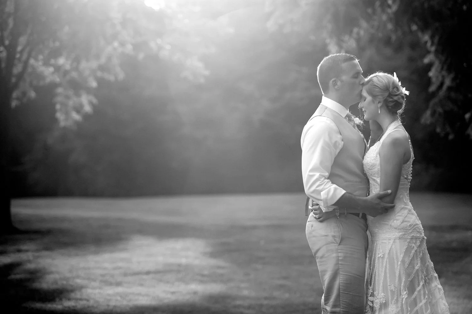 A black and white photo of a bride and groom sharing a kiss outdoors on a grassy field with trees in the background, illuminated by soft sunlight.