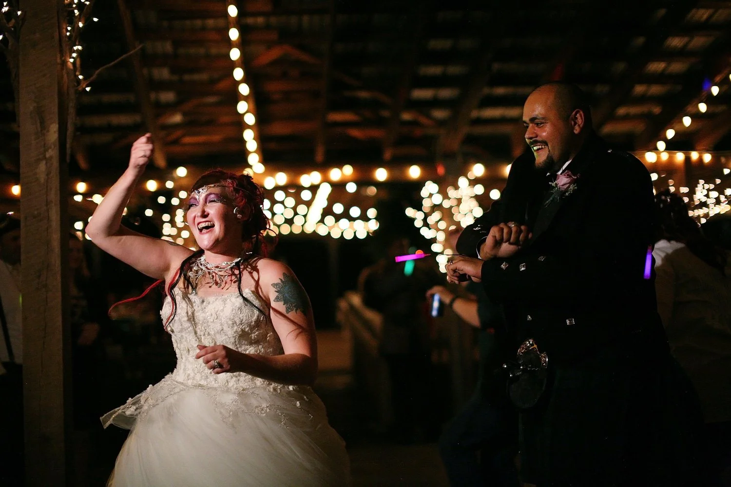 A bride and groom dancing and celebrating at a wedding reception with string lights overhead and ambient lighting. Her dress has tons of tulle and her arms are up above her as she dances.