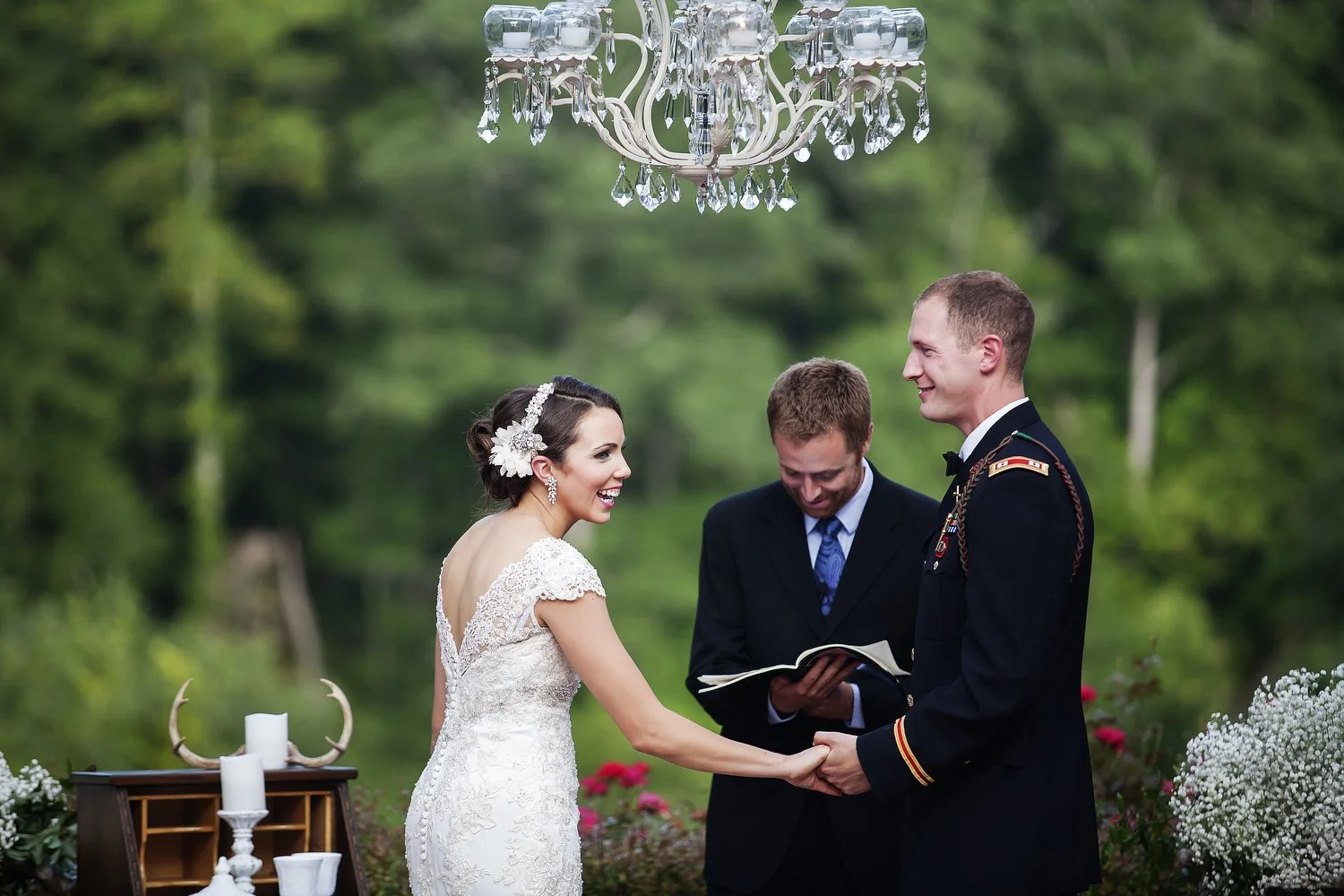 A bride and groom hold hands during their outdoor wedding ceremony, smiling at each other. An officiant stands between them with a book, in a lush green garden with bouquet and decorative candles on a table nearby, and a chandelier hanging overhead.