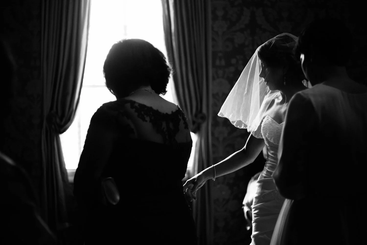 A black and white photo of a bride in a wedding dress and veil over her face. She is surrounded by two women in front of a window with curtains and natural light coming in the room. The veil has gorgeous window light making it stand out against the d