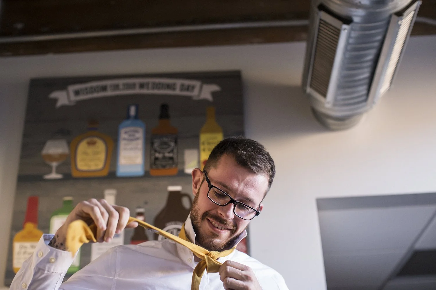 A man with glasses and a beard, ties his yellow tie in a well-lit room. There is an industrial pipe on the ceiling behind him and a huge wooden artwork with glasses and alcohol bottles painted on it.