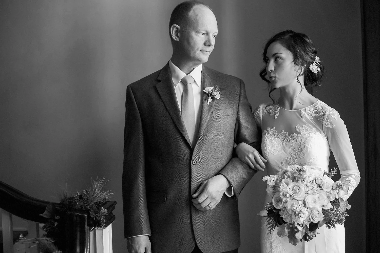 Black and white photo of a bride and a man, possibly her father, before a wedding, with the bride holding a bouquet of flowers and wearing a lace wedding dress, while the man is in a suit with a boutonniere.