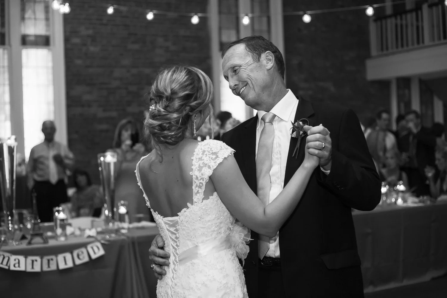 A bride and her dad dancing at the wedding reception. The bride wears a white lace dress with a low back, and her dad is dressed in a black tuxedo with a white shirt and a light-colored tie. The background has decorations and guests at tables.