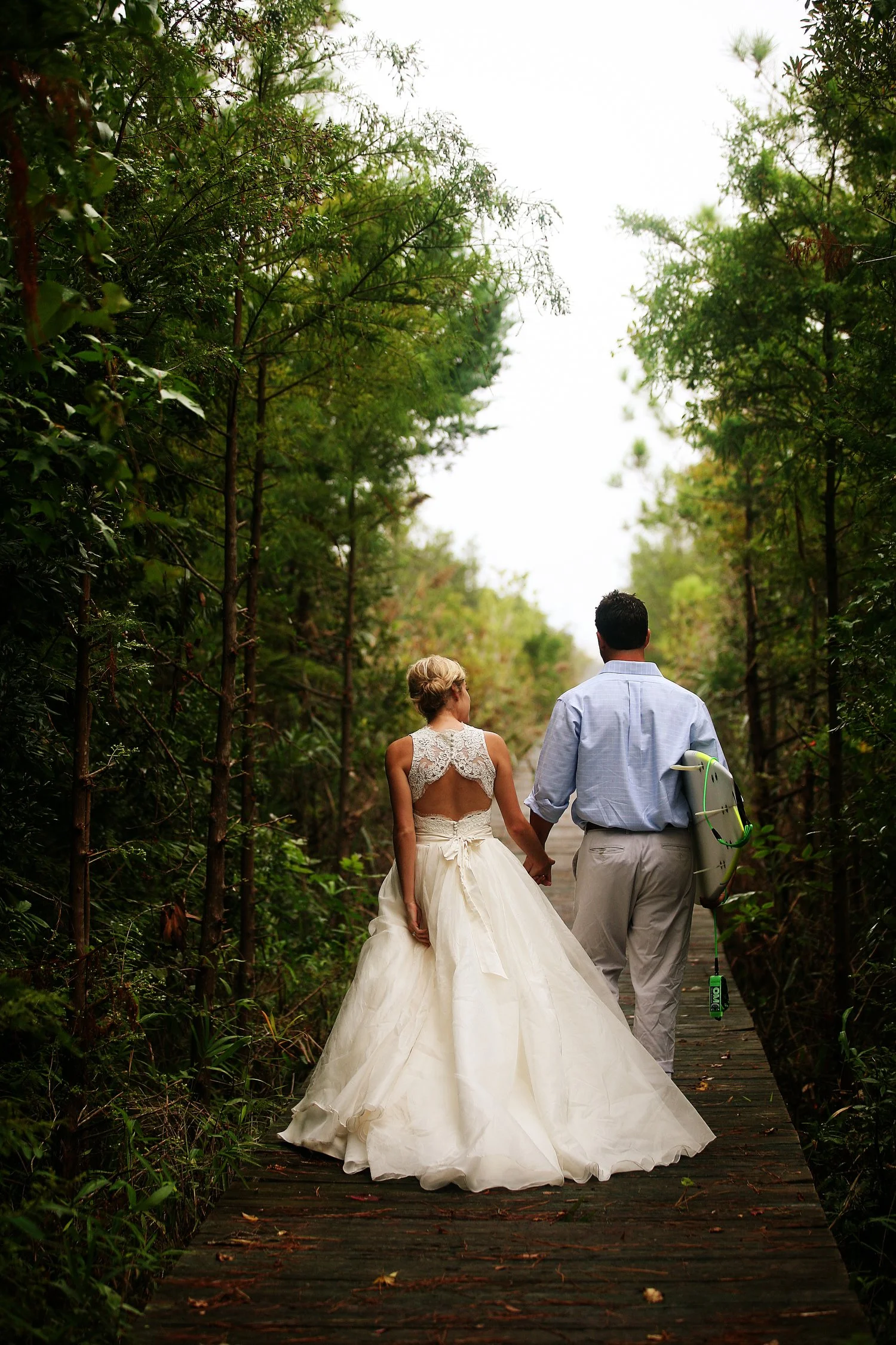 A bride and groom walking hand in hand on a wooden path through a lush green forest. The bride is wearing a white wedding gown, and the groom is dressed in a light blue shirt and light-colored trousers, holding a surfboard.