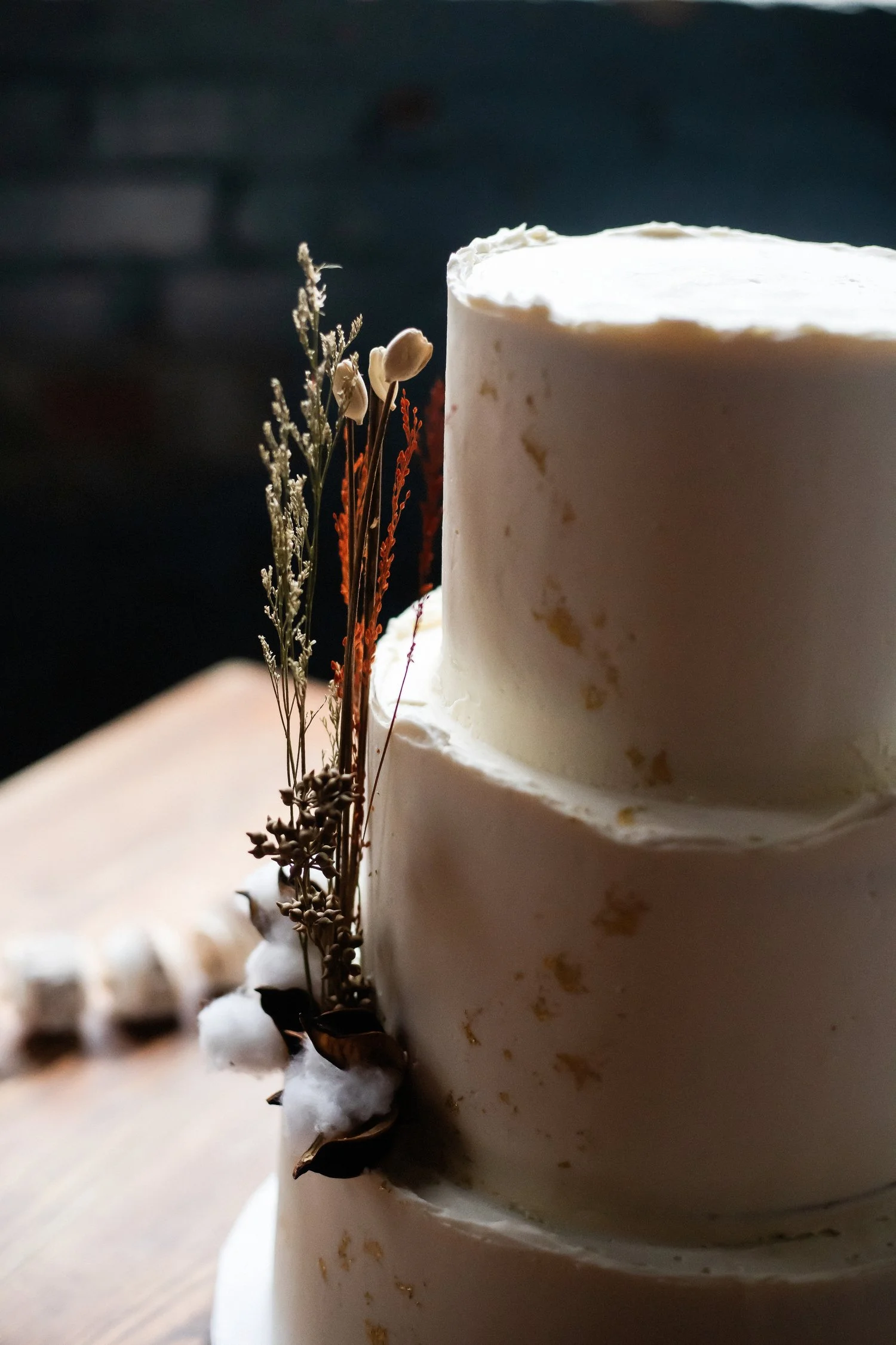The top two tiers of a round three-tier cake are visible close up. The cream-colored frosting has some gold flecks in it. The bottom layer is adorned with some cotton and pieces of wheat-like plants. 