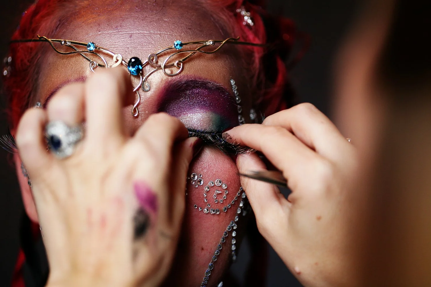 Close up shot of hands applying false eyelashes onto a woman's face. The woman has bright reddish-pink hair and purple eyeshadow. She has a jewel crown around her forehead and other pieces of jewels pressed onto her face.