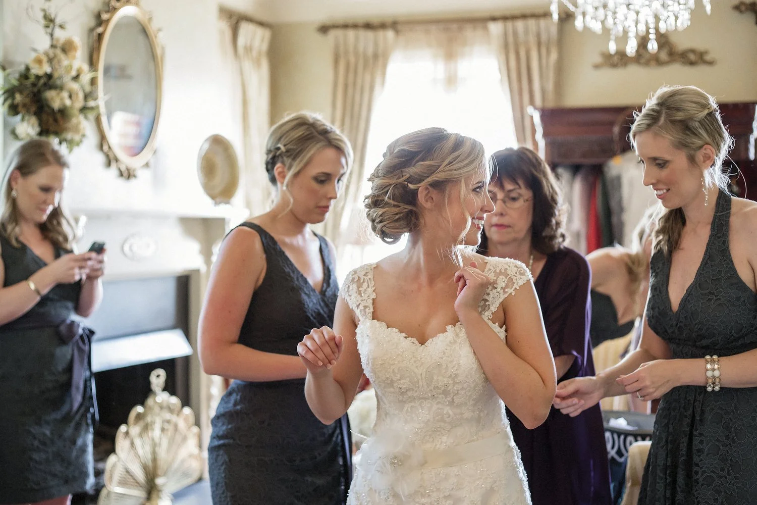 Bridesmaids and family help a bride with her dress in a vintage decorated room before the wedding ceremony. The bride has on a white lacy dress with lacy shoulder straps, and her hair is in a loose updo with wavy tendrils falling next to her face.