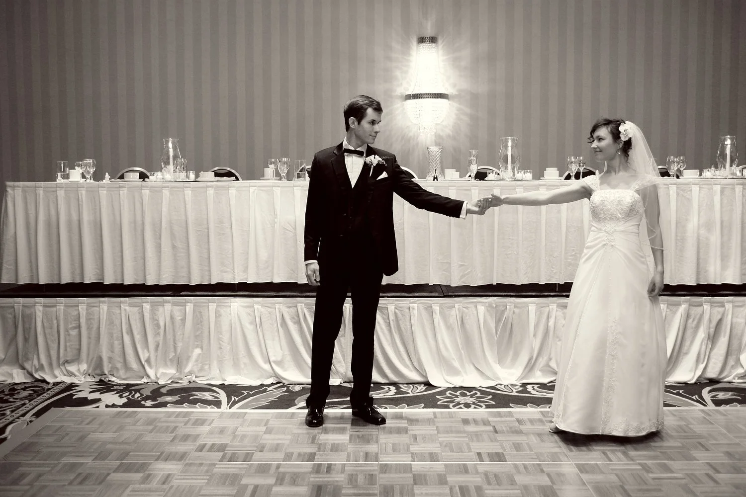 A bride and groom stand apart with arms outstretched holding hands on the dance floor at their wedding reception preparing for their first dance. He is wearing a black suit and a bowtie. She is in a white wedding dress with a veil. There is a long di