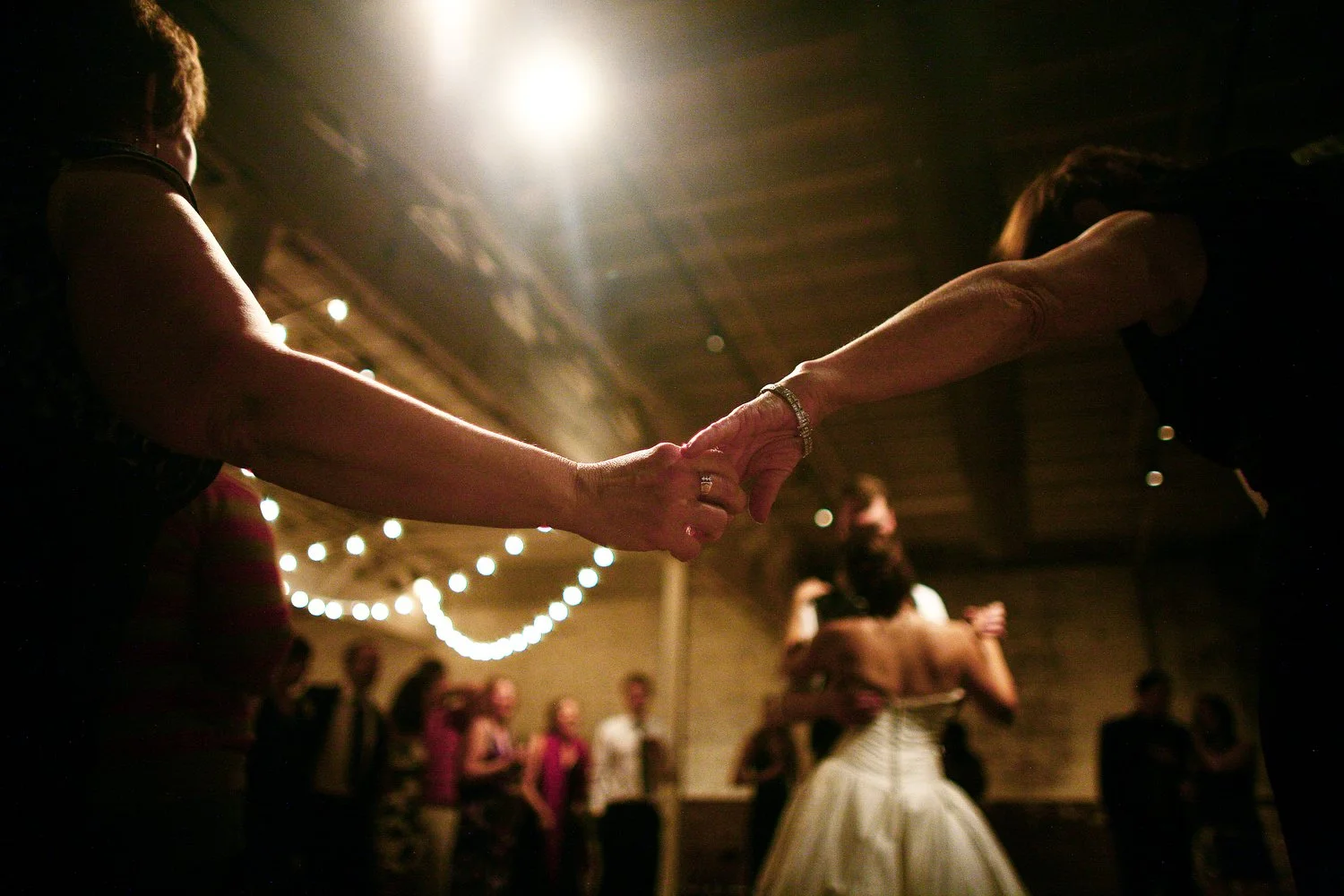 The silhouettes of two women's arms outstretched holding hands frame the forefront of the image. Underneath the clasped hands, and blurred in the distance, is a bride and groom dancing. She has her left arm on his shoulder and holds his hand with her