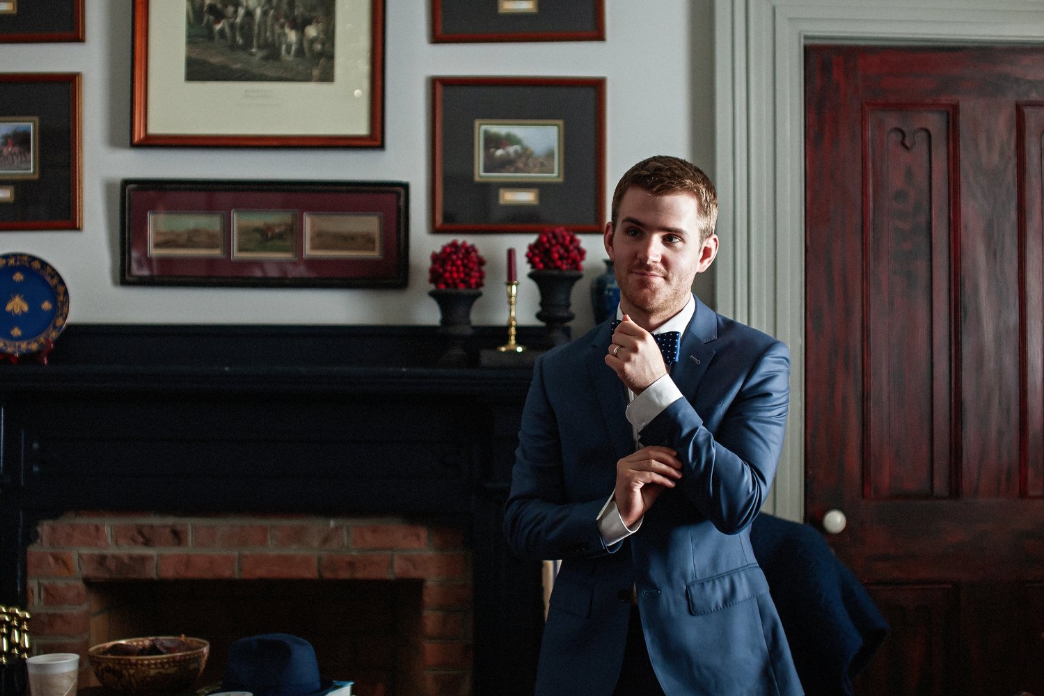 A man in a blue suit with a white shirt and bow tie standing indoors with a thoughtful expression, in front of a wall with framed pictures and a fireplace. He is adjusting the cuff of his sleeve.