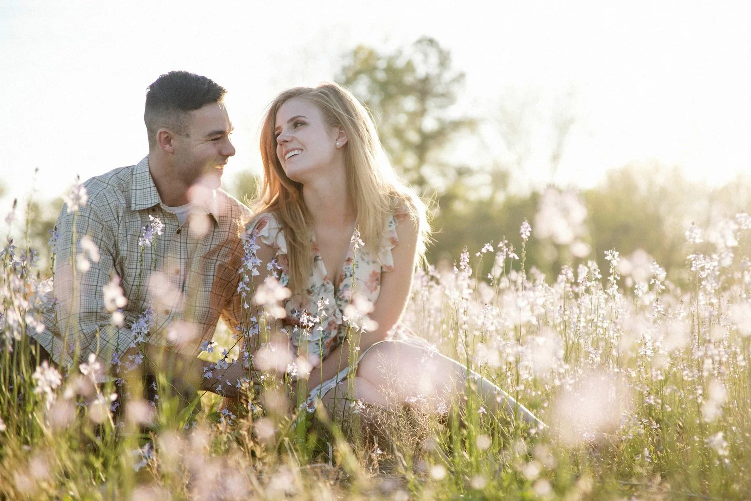 Warm sunlight serves as beautiful backlighting for a couple sitting in a field with tiny purple flowers blurred intentionally in the foreground. They are smiling and looking at each other. Her long blond hair is lit by the sun and she is wearing some