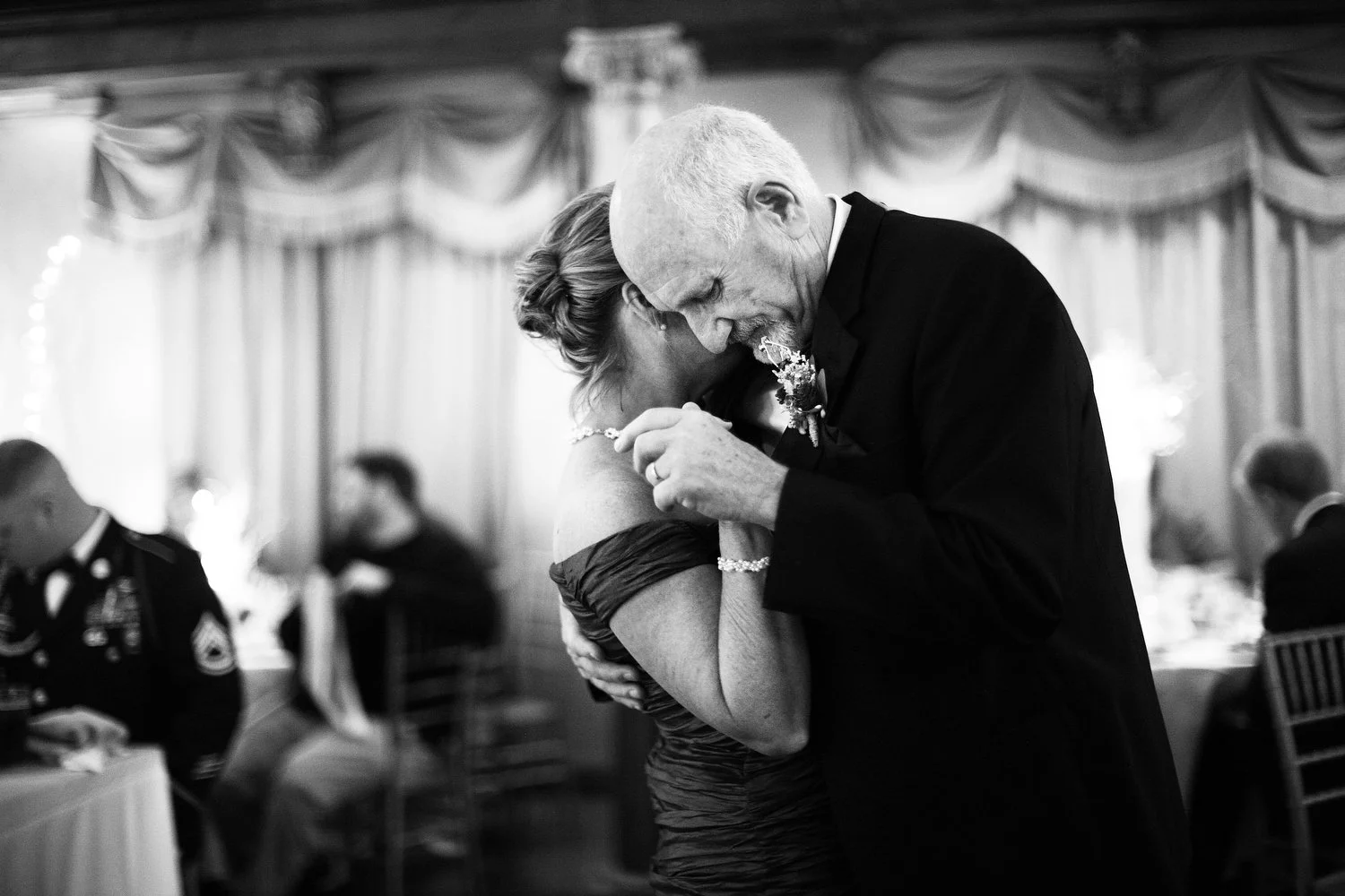 An elderly man and woman dancing closely at a formal event, both with their eyes closed and smiling, in a room with curtains and other guests seated at tables in the background.