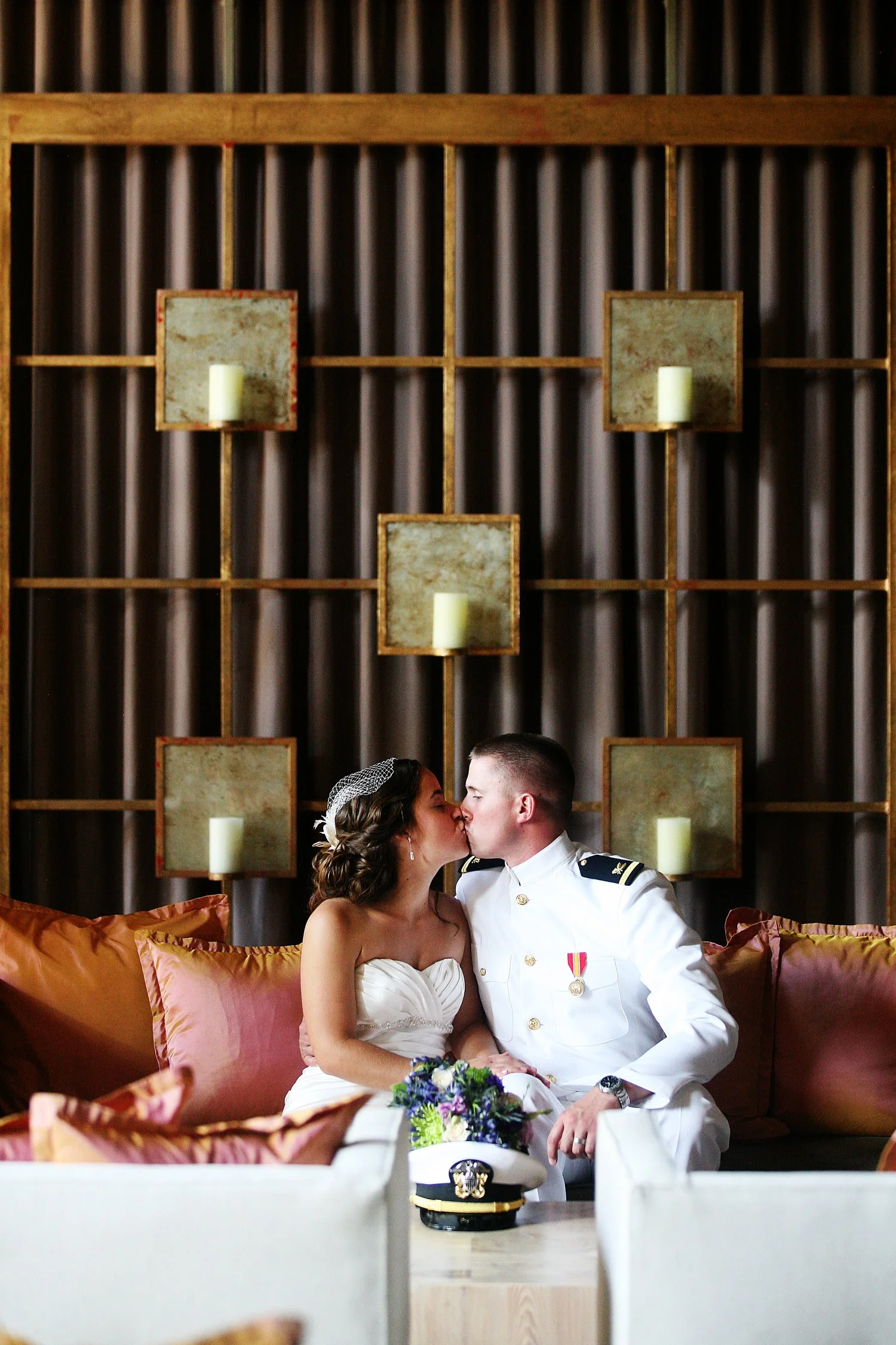 A bride and groom in wedding attire sharing a kiss in an indoor setting with modern decor and candles on the wall behind them. He is in Navy dress whites.