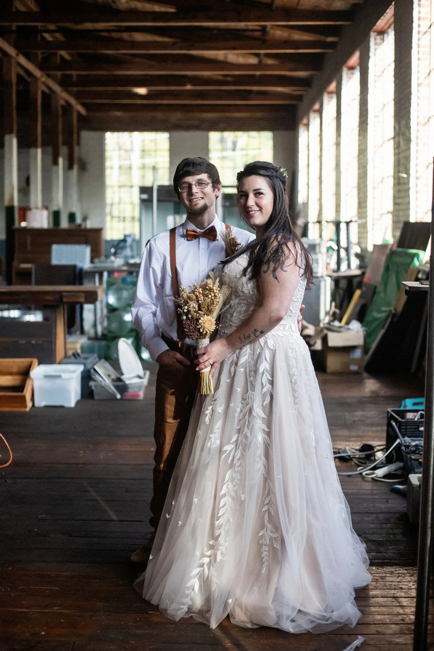 A bride and groom standing together indoors, with the bride holding a bouquet of dried flowers, in a rustic space with large windows and wooden beams.