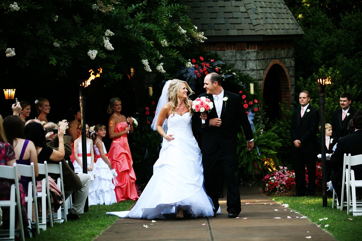 A bride and groom walking down the aisle during their wedding ceremony outside, surrounded by guests, bridesmaids, and groomsmen, with floral arrangements and tiki torches.