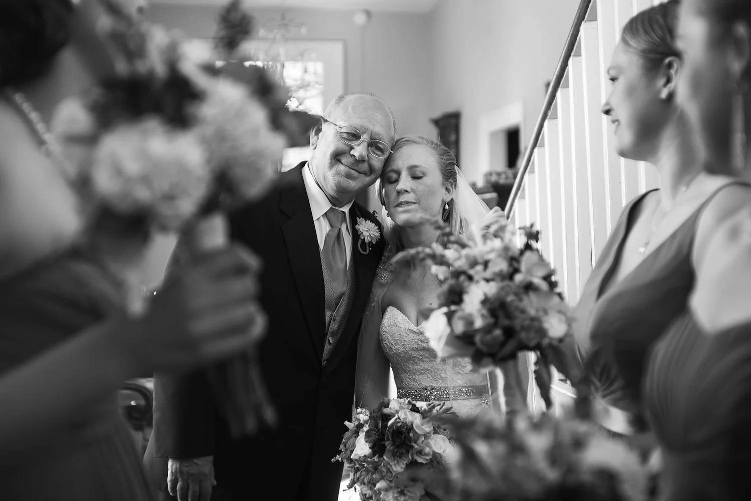 Black and white photo of a bride with her eyes closed, leaning on a man likely her father, who is smiling with glasses. They are surrounded by bridesmaids holding bouquets, smiling and looking at the bride in the foreground.