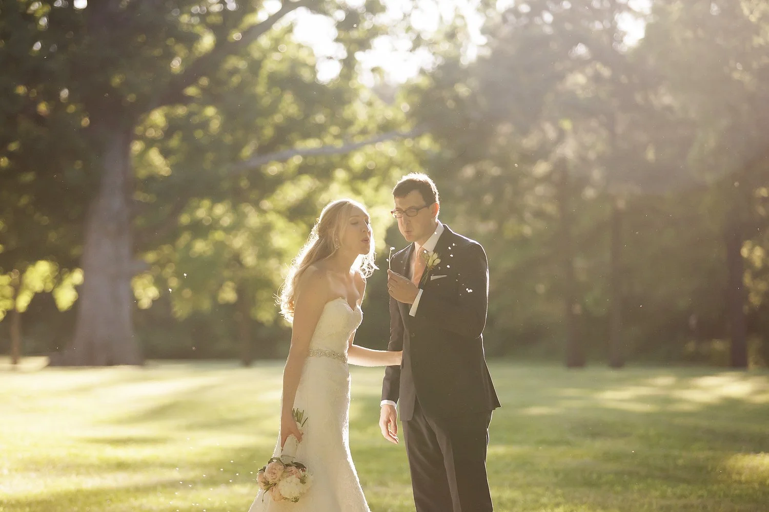 A bride and groom standing outdoors, illuminated by sunlight, with trees in the background blowing a dandelion in sunset light.