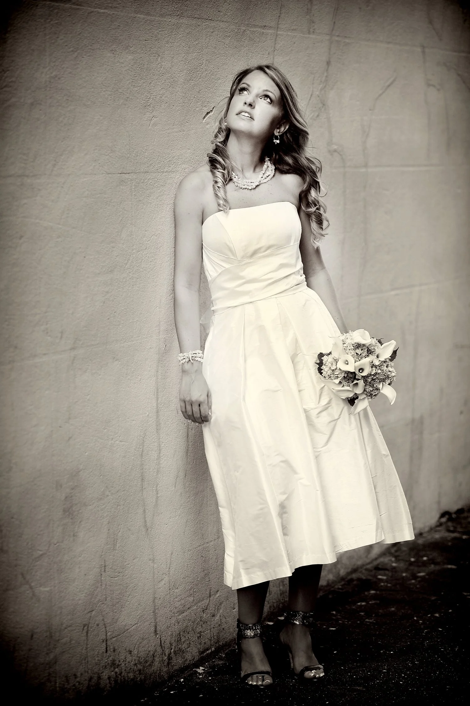 Black and white portrait of a bride leaning against a white concrete wall and looking up and off in the distance. She is wearing a tea length strapless white dress. She has on high heels with straps around her ankles and has on a necklace and bracele