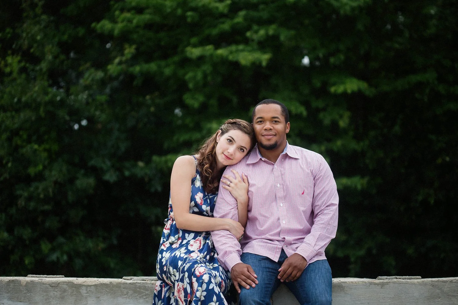 A couple sits on a low concrete wall with lush green trees behind them. She is wearing a sleeveless floral dress and has her arms wrapped around his arm as she rests her head on his shoulder. She has a soft smile on her face. He is wearing a pink dre