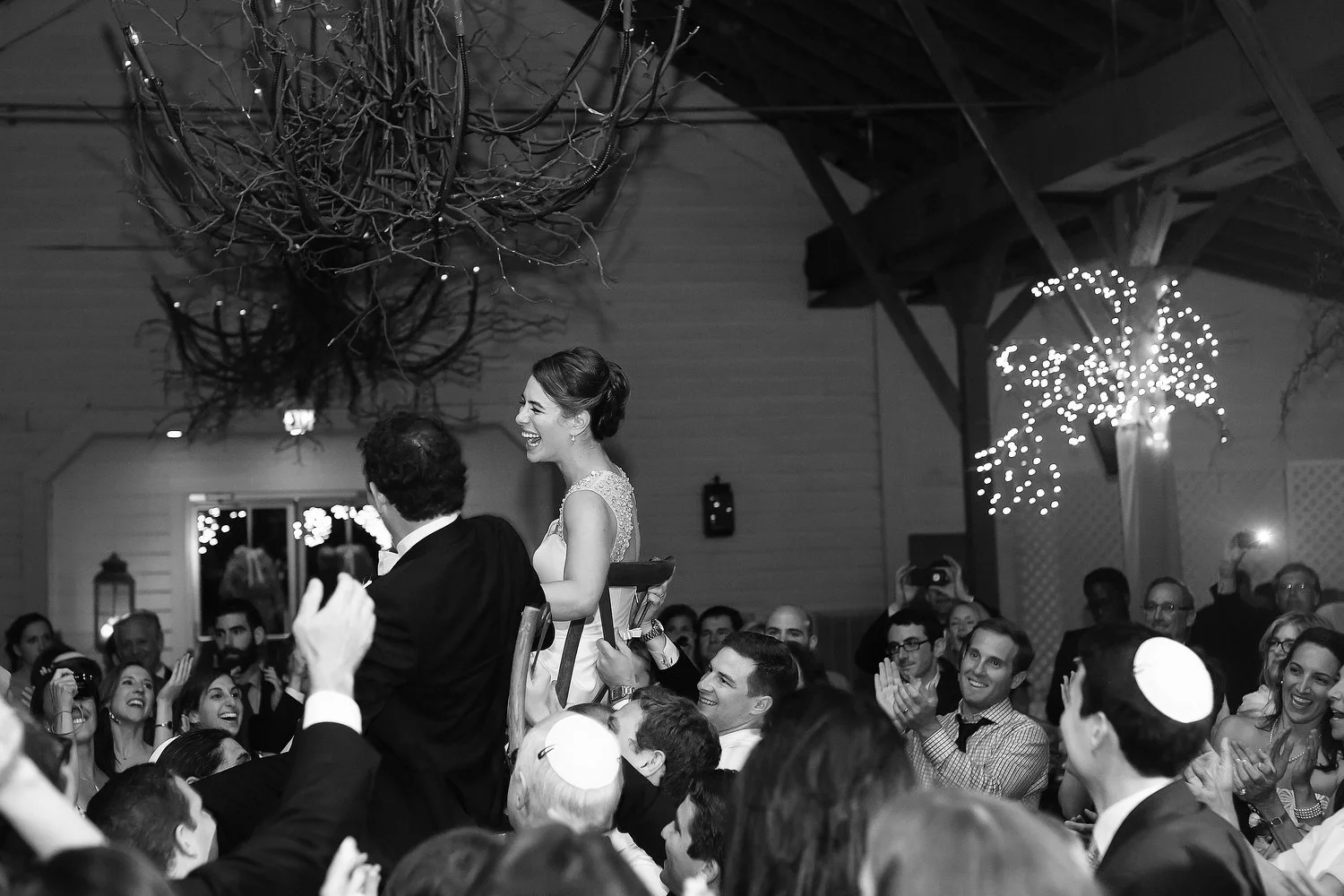 A joyful bride is lifted on a chair during a celebration, surrounded by smiling guests at a wedding reception in a decorated venue. This is the Jewish hora dance tradition.