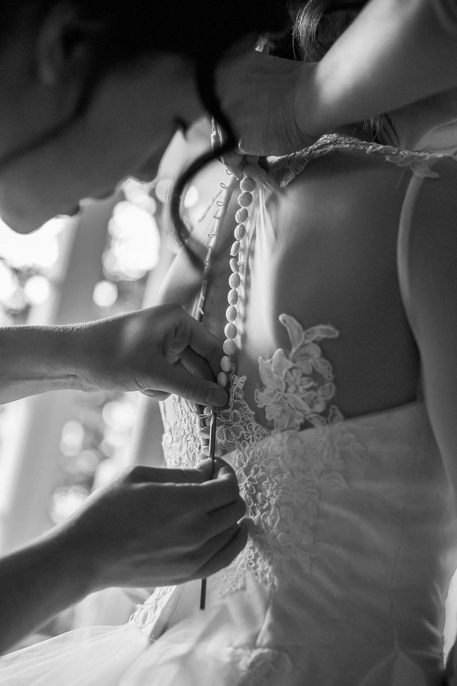 Close-up of someone adjusting the back of a bride's wedding dress, focusing on detailed lace and fabric, in black and white. The woman is using a crochet hook to secure the loops around each button.