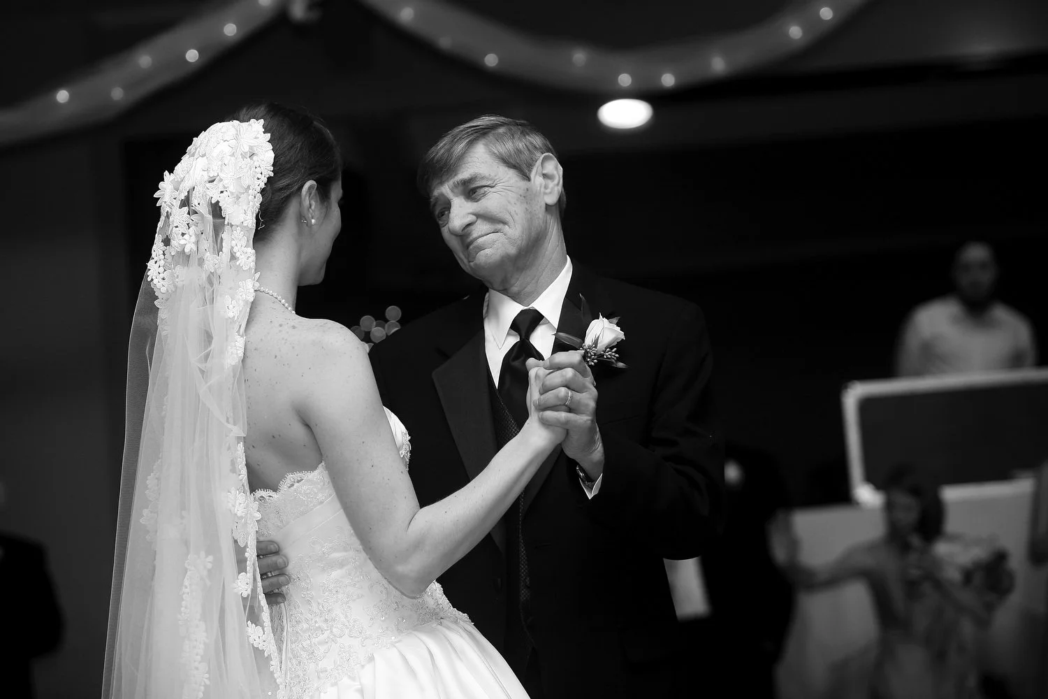 An older man with dark grey hair smiles tenderly as he looks at his daughter in her wedding dress. They are holding hands and dancing the father-daughter dance at her wedding reception. She has on a strapless white gown and a chapel length veil. The 